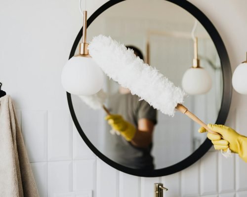 A person cleaning an elegant bathroom mirror with a fluffy duster and yellow gloves.